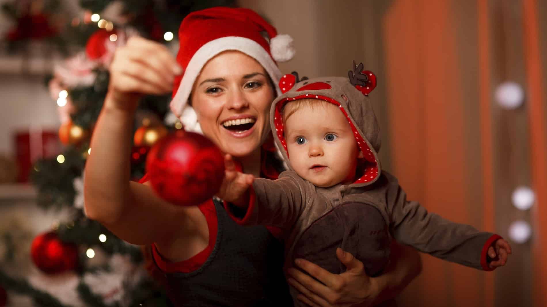 Happy mother showing Christmas ball to baby near Christmas tree.